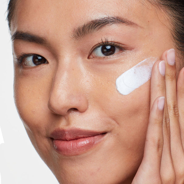 Close-up of a woman gently applying Solar Defense Non-Tinted SPF50 | HydroPeptide sunscreen to her cheek, showcasing smooth skin and skincare routine.