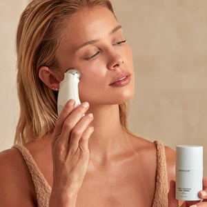Woman using a facial device on her cheek while holding a bottle of Firming + Brightening Silk Crème | NuFACE skincare product in a neutral-toned setting