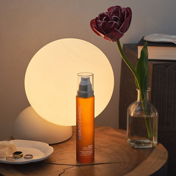 Night Body Serum | Moroccanoil in an amber bottle with a pump top, displayed on a wooden table next to a glowing round lamp and a purple tulip in a glass vase, creating a cozy skincare scene.
