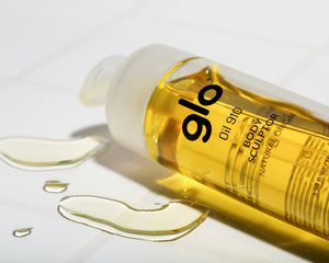 Close-up of a bottle of Oil910 | Glo910 body sculptor oil with spilled golden liquid on a white surface, highlighting a natural origin skincare product for wellness and body care.