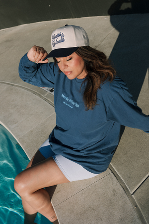 Woman sitting by a pool wearing a two-tone cap with "Health Is Wealth Hat | Lucky Owl" embroidered on the front, paired with a blue long-sleeve shirt and white shorts.