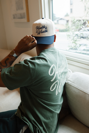 Person wearing a white and blue cap with "Health Is Wealth Hat | Lucky Owl" embroidered text, sitting on a couch near a window, wearing a green shirt with matching white script text on the back.