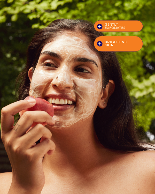 Woman with a glowing smile and exfoliating mask on her face holding a fruit, promoting the Superfruit Lactic + Multifruit 8% AHA Exfoliating Mask | Three Ships for skincare and brightening benefits.