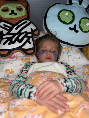 Child resting peacefully with eye patches on, lying in bed surrounded by two large Spa Bunny Plushie | Lucky Owl cushions featuring playful facial designs.