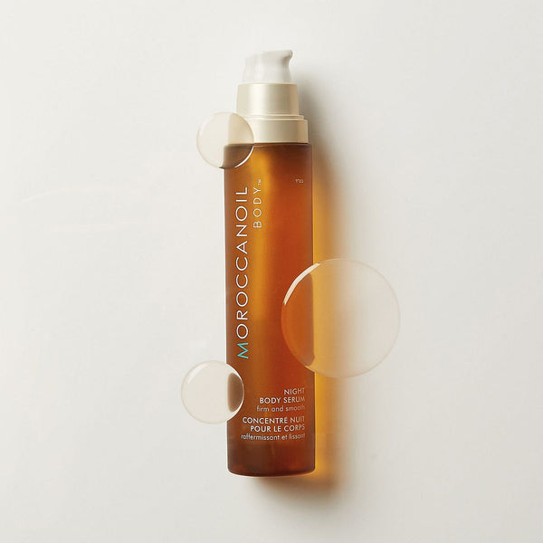 Bottle of Night Body Serum | Moroccanoil in a sleek amber pump container surrounded by clear liquid droplets, highlighting skincare and body care wellness.