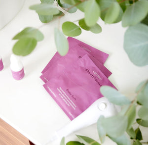 Three pink packets of LightStim SheetMasque | LightStim skincare product arranged on a white surface surrounded by green leaves and a white beauty device partially visible.