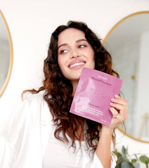 Young woman with curly hair smiling and holding a pink packet of LightStim SheetMasque | LightStim skincare product in a bright bathroom setting with round mirrors and plants in the background