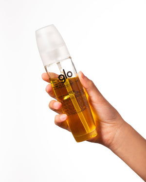 Hand holding a clear bottle filled with golden oil featuring the label "glo" against a white background, representing the Oil910 | Glo910 skincare product.