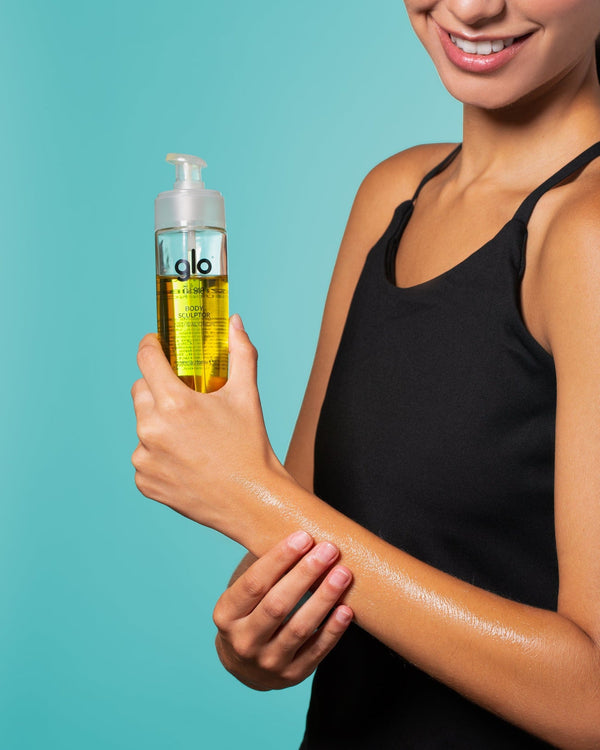 Smiling woman wearing a black tank top holding a bottle of Oil910 | Glo910 skincare oil while applying it to her glossy forearm against a turquoise background