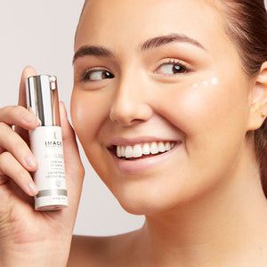 Smiling woman holding a bottle of AGELESS total eye lift crème | IMAGE Skincare with small dots of the crème applied under her eye in a bright skincare setting.