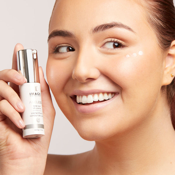 Smiling woman holding a bottle of AGELESS total eye lift crème | IMAGE Skincare with small dots of the crème applied under her eye in a bright skincare setting.