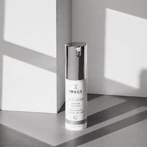 Bottle of AGELESS total eye lift crème | IMAGE Skincare with reflective silver cap, set against a minimalist gray and white background with soft natural light and shadows.