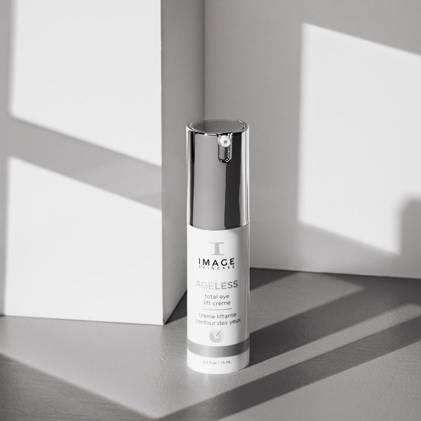 Bottle of AGELESS total eye lift crème | IMAGE Skincare with reflective silver cap, set against a minimalist gray and white background with soft natural light and shadows.
