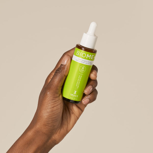 Hand holding a green bottle of BIOME+ dew bright serum | IMAGE Skincare against a plain beige background, highlighting skincare and wellness.
