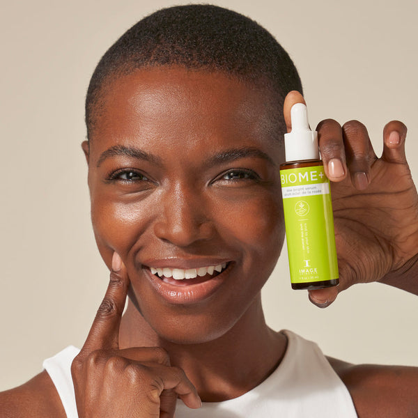 Smiling woman with smooth skin holding a green bottle of BIOME+ dew bright serum | IMAGE Skincare near her face, showcasing skincare and wellness.