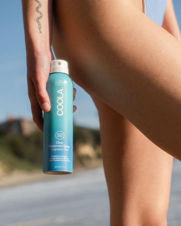 Person holding Classic Body Organic Sunscreen Spray SPF 50 - 6 fl oz | COOLA by their thigh at the beach, showcasing skincare product for sun protection and body care.