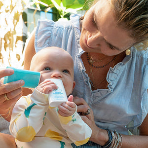 A smiling woman applies Mineral Stick SPF 50 | COOLA sunscreen to a baby holding the product, showcasing gentle skincare and sun protection for sensitive skin outdoors.