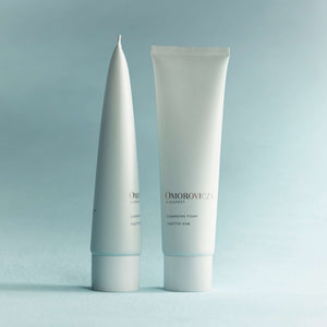 Two sleek white tubes of Cleansing Foam | Omorovicza standing upright against a soft blue background, showcasing minimalist skincare packaging design.