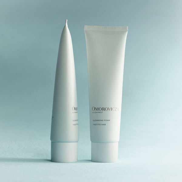 Two sleek white tubes of Cleansing Foam | Omorovicza standing upright against a soft blue background, showcasing minimalist skincare packaging design.