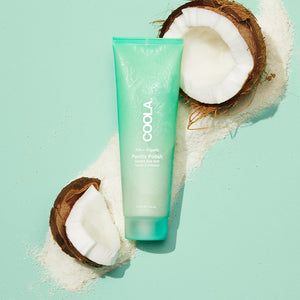 Pacific Polish Gentle Sea Salt Facial Exfoliator | COOLA tube displayed on a light turquoise background with natural sea salt and cracked coconuts, highlighting skincare wellness.