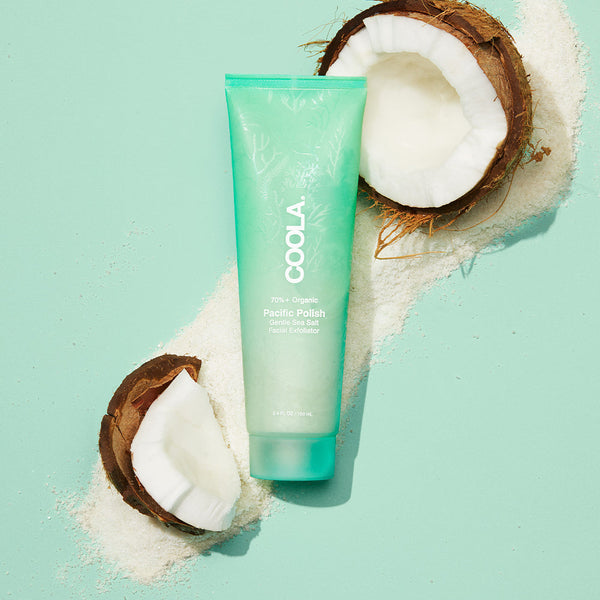 Pacific Polish Gentle Sea Salt Facial Exfoliator | COOLA tube displayed on a light turquoise background with natural sea salt and cracked coconuts, highlighting skincare wellness.