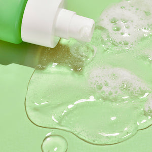 Close-up of clear foaming gel dispensed from a green bottle pump on a light green surface, showcasing the texture of Glowing Greens Detoxifying Facial Cleansing Gel | COOLA skincare product.