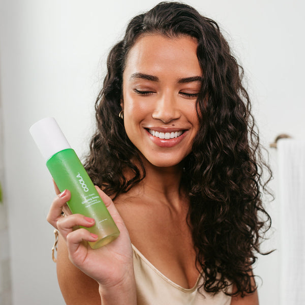 A smiling woman with curly hair holding a green bottle of Glowing Greens Detoxifying Facial Cleansing Gel | COOLA, showcasing skincare in a bright, clean setting.