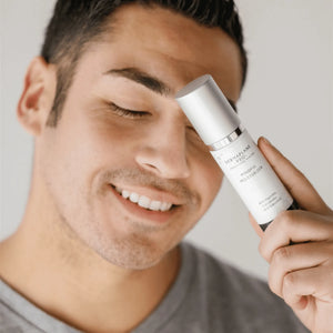 Smiling man holding a white bottle of Mindful Moisturizer | DermaplanePro skincare product near his face, promoting wellness and hydration.