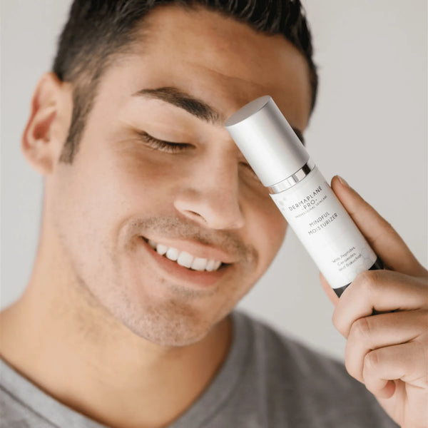 Smiling man holding a white bottle of Mindful Moisturizer | DermaplanePro skincare product near his face, promoting wellness and hydration.