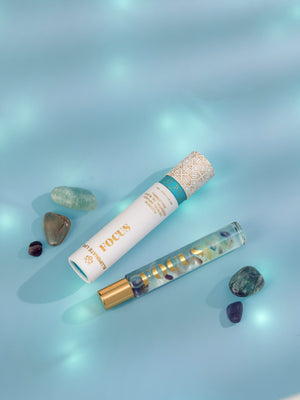 Focus Rollerball | Aluminate Life with a decorative white and gold cap next to a glass rollerball bottle filled with colorful stones, surrounded by polished crystals on a soft blue surface.