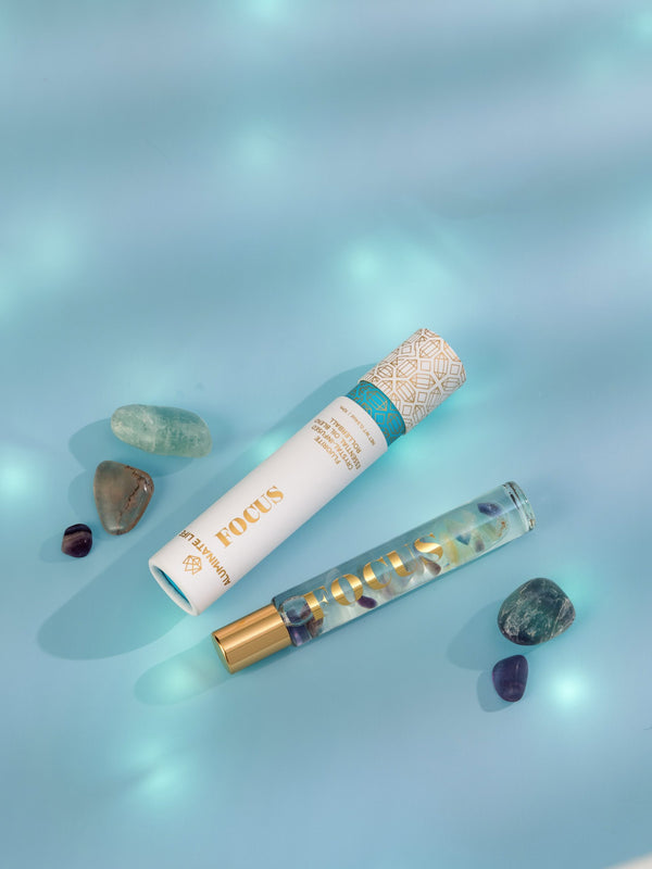 Focus Rollerball | Aluminate Life with a decorative white and gold cap next to a glass rollerball bottle filled with colorful stones, surrounded by polished crystals on a soft blue surface.
