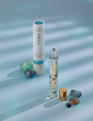Focus Rollerball | Aluminate Life featuring a clear rollerball bottle with colorful stones inside, paired with its decorative white and blue cylindrical packaging and surrounding polished crystals.