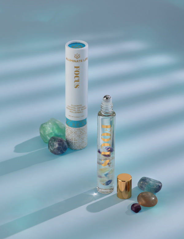 Focus Rollerball | Aluminate Life featuring a clear rollerball bottle with colorful stones inside, paired with its decorative white and blue cylindrical packaging and surrounding polished crystals.