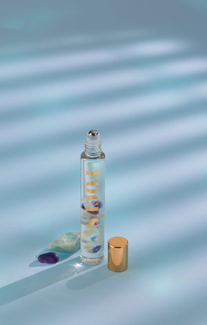 Focus Rollerball | Aluminate Life wellness tool featuring a clear glass bottle with colorful stones inside and a gold cap, set against a soft blue background with light shadows.