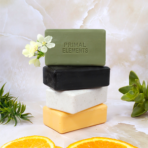 Stack of four assorted natural soap bars with the top green bar embossed "PRIMAL ELEMENTS" surrounded by small white flowers and fresh greenery, highlighting Luxury Shea Butter Moisturizing Soap | Primal Elements for skincare and body care.