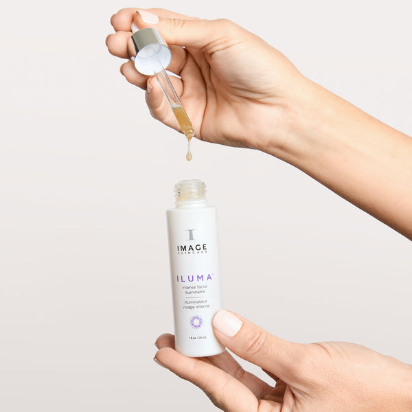 Hands holding a white bottle and dropper of ILUMA intense facial illuminator | IMAGE Skincare with a drop of serum about to fall from the dropper, skincare product demonstration.