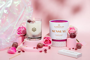 Sensual Glass Candle | Aluminate Life with lit wick, surrounded by pink roses, crystal stones, matches, and a branded box on a soft pink background creating a calming wellness scene.