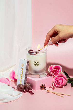 Hand lighting a Sensual Glass Candle | Aluminate Life surrounded by pink roses, rose petals, gemstone crystals, and a branded matchbox on a soft pink background.