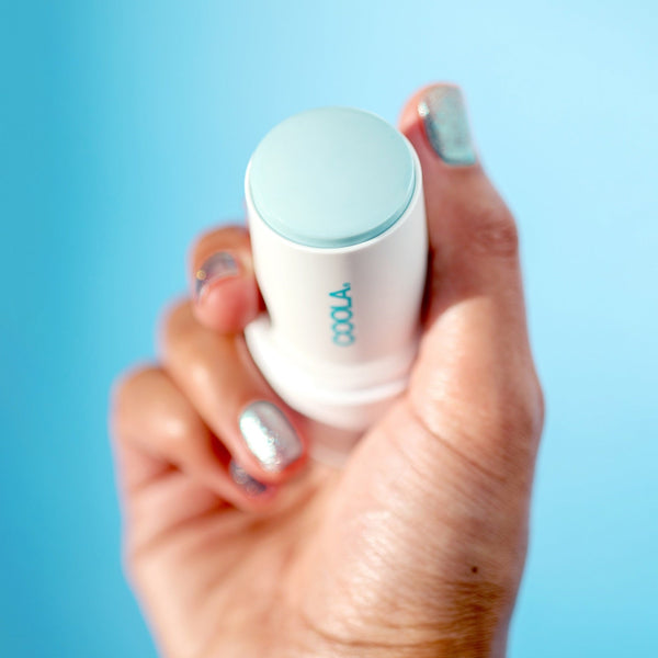 Hand holding a light blue and white Refreshing Water Stick SPF 50 | COOLA against a blue background, showcasing skincare product with silver glitter nail polish on fingers.