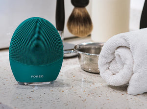 LUNA™ 4 MEN | FOREO facial cleansing brush in teal displayed on a bathroom counter next to a rolled white towel and shaving brush for skincare and grooming routine.