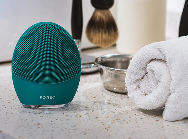 LUNA™ 4 MEN | FOREO facial cleansing brush in teal displayed on a bathroom counter next to a rolled white towel and shaving brush for skincare and grooming routine.