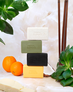 Four stacked bars of Luxury Shea Butter Moisturizing Soap | Primal Elements in white, green, black, and yellow colors next to fresh oranges and green leaves on a marble surface.