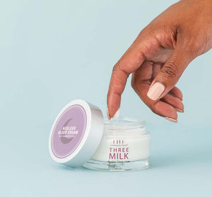 Hand dipping finger into an open jar of Three Milk Ageless Sleep Cream on a blue background, representing skincare from the Silky, Milky & Most Loved Naked Nourishment Set | Farmhouse Fresh.