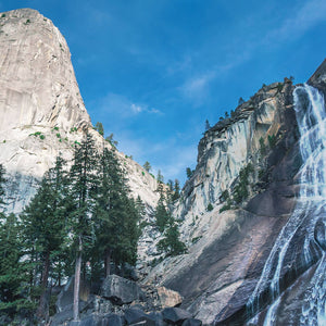 Scenic view of Yosemite National Park featuring towering granite cliffs, lush pine trees, and a cascading waterfall under a clear blue sky, evoking the essence of the Yosemite 8oz Candle | The Beauty of Hope.