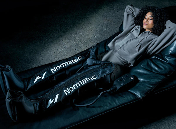 A woman relaxing on a black leather chaise lounge using the Normatec 3 Leg Recovery System | Hyperice on her legs, demonstrating a recovery tool for wellness and body care.