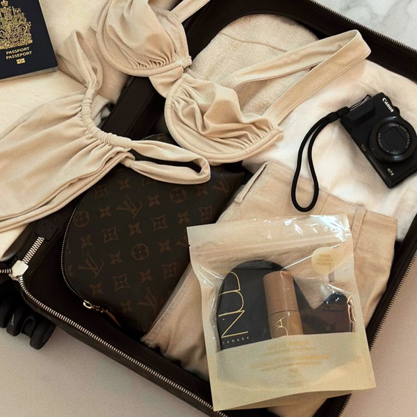 Open suitcase packed with beige clothing, a passport, a camera, a designer bag, and the Self Tan Travel Kit | NUDA in a clear resealable pouch for skincare on the go.