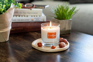 I Choose Optimism Candle | Aluminate Life lit on a wooden tray surrounded by polished stones, with plants and books softly blurred in the background, creating a calm wellness scene.