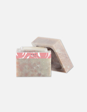 Two bars of natural soap with a soft earthy tone and a pink label, next to a clear lip balm, representing Concreta | Zents skincare products.