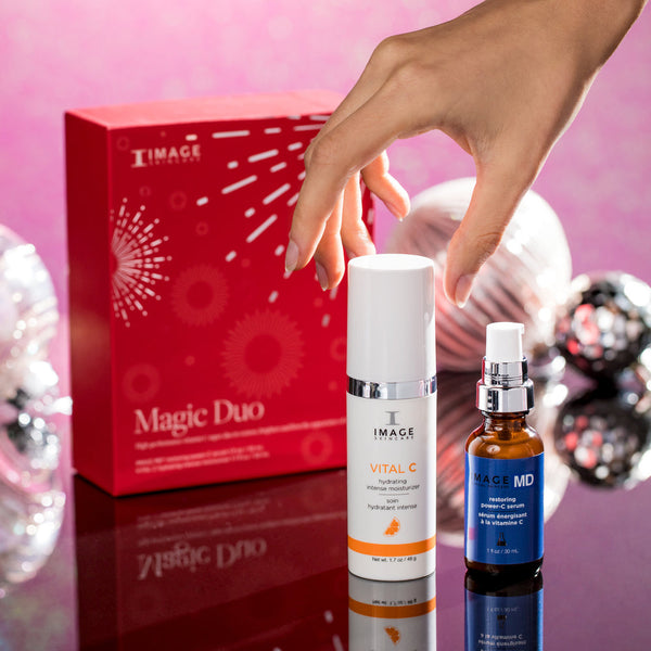 Hand reaching for skincare products including a white bottle and a blue dropper bottle in front of a red Magic Duo | IMAGE Skincare box on a reflective surface with festive decorations in the background.