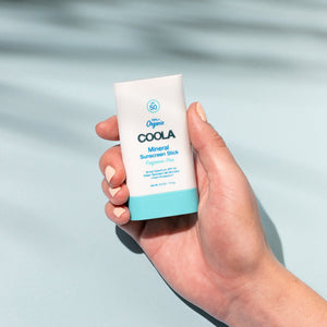 Hand holding a Mineral Stick SPF 50 | COOLA sunscreen stick with a light blue and white design, set against a soft pale blue background with subtle shadows.